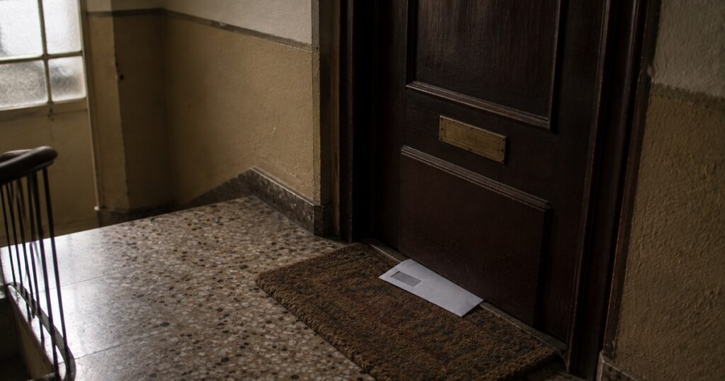 Rellano con la puerta de un piso cerrada y un sobre asomando por debajo