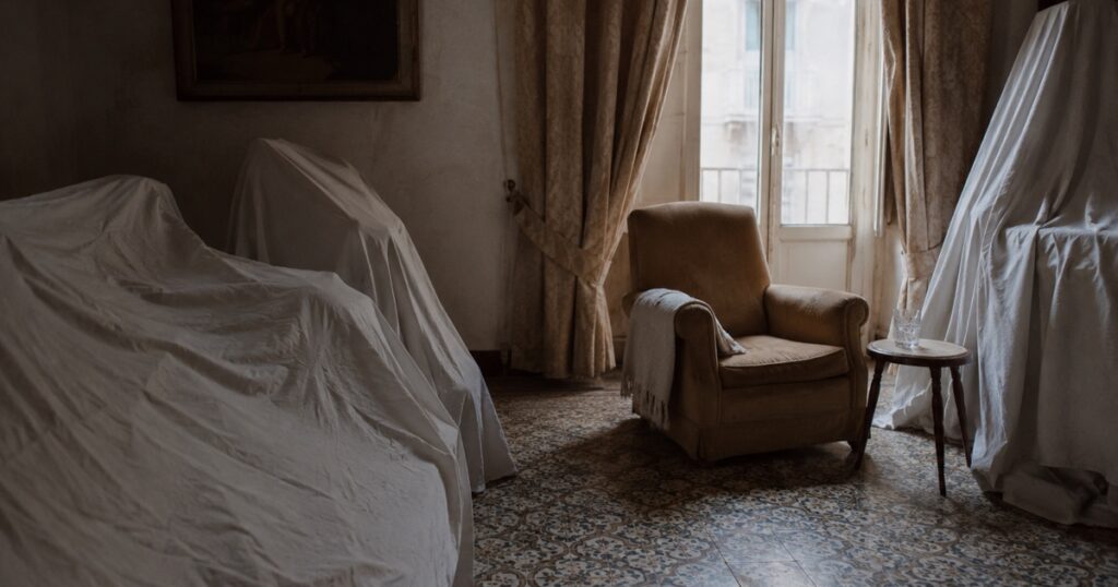 Un sillón junto a la ventana y el resto de los muebles cubiertos con sábanas