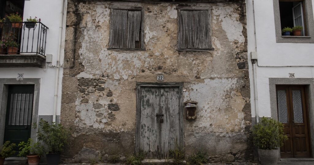 Casa de pueblo cerrada y con maleza en la puerta, entre dos viviendas habitadas